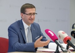 ein Mann im Anzug spricht vor einem Pult mit vielen Mikrofonen.