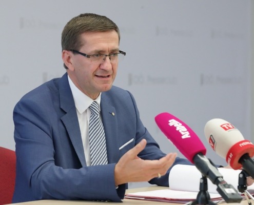 ein Mann im Anzug spricht vor einem Pult mit vielen Mikrofonen.