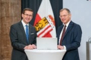 Zwei Männer in Anzügen stehen an einem runden Tisch mit Laptop, lächeln in die Kamera; im Hintergrund Fahnen eines österreichischen Bundeslandes