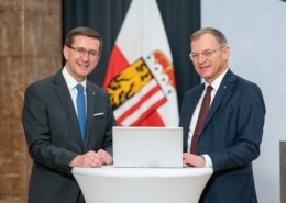 Zwei Männer in Anzügen stehen an einem runden Tisch mit Laptop, lächeln in die Kamera; im Hintergrund Fahnen eines österreichischen Bundeslandes