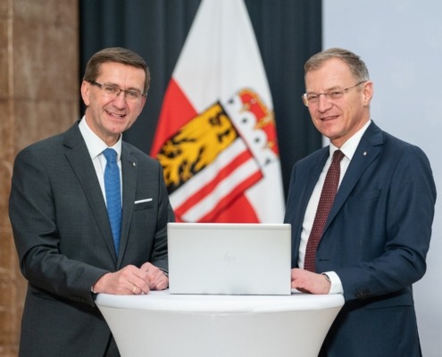 Zwei Männer in Anzügen stehen an einem runden Tisch mit Laptop, lächeln in die Kamera; im Hintergrund Fahnen eines österreichischen Bundeslandes