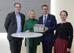 Vier Menschen stehen von einem Stehtisch und lächeln in die Kamera. Ein Herr hält eine Kiste in der Hand.