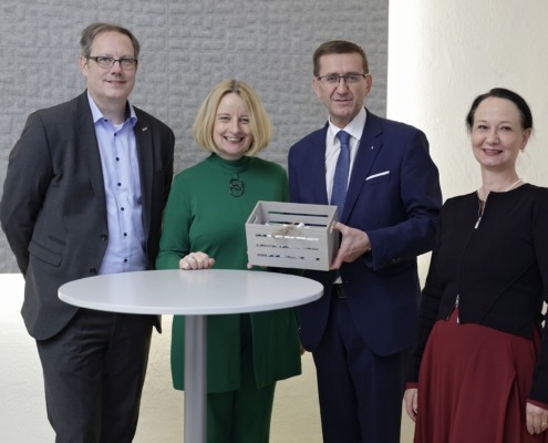 Vier Menschen stehen von einem Stehtisch und lächeln in die Kamera. Ein Herr hält eine Kiste in der Hand.