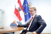 LR_Achleitner_3_Land_OOE_Liedl Ein Herr sitz an einem Tisch und redet. Hinter ihm sind 2 Flaggen. Eine Österreichflagge und eine Europaflagge.