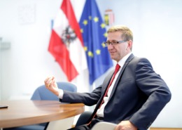 Ein Herr sitz an einem Tisch und redet. Hinter ihm sind 2 Flaggen. Eine Österreichflagge und eine Europaflagge.