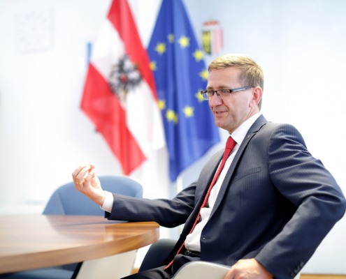 Ein Herr sitz an einem Tisch und redet. Hinter ihm sind 2 Flaggen. Eine Österreichflagge und eine Europaflagge.