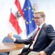 Ein Herr sitz an einem Tisch und redet. Hinter ihm sind 2 Flaggen. Eine Österreichflagge und eine Europaflagge.