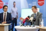 Zwei Männer in Anzügen bei einer Pressekonferenz: Einer spricht am Stehtisch in mehrere Mikrofone, daneben ein Kollege; im Hintergrund Werbewand und unscharfe Person.