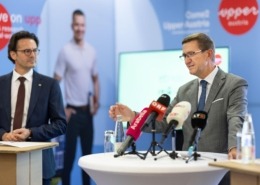 Zwei Männer in Anzügen bei einer Pressekonferenz: Einer spricht am Stehtisch in mehrere Mikrofone, daneben ein Kollege; im Hintergrund Werbewand und unscharfe Person.