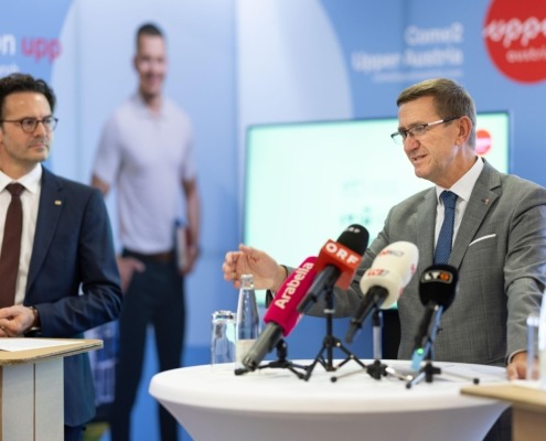 Zwei Männer in Anzügen bei einer Pressekonferenz: Einer spricht am Stehtisch in mehrere Mikrofone, daneben ein Kollege; im Hintergrund Werbewand und unscharfe Person.