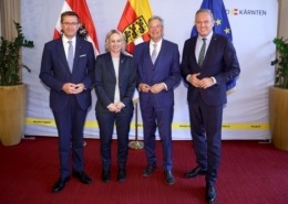 Vier Herren stehen vor einer Wand. Vor der Wand sind 3 Flaggen. Die Herren tragen Anzüge und lächeln in die Kamera.