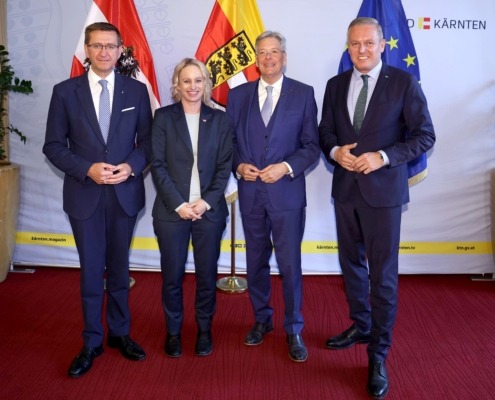 Vier Herren stehen vor einer Wand. Vor der Wand sind 3 Flaggen. Die Herren tragen Anzüge und lächeln in die Kamera.