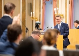 Ein Mann im Anzug steht am Rednerpult in einem Parlamentssaal und spricht. Vor ihm sitzen Zuhörer, im Hintergrund Kameras und Wandlampen an den Wänden.