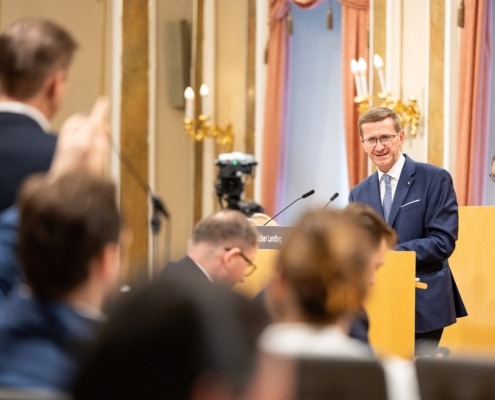 Ein Mann im Anzug steht am Rednerpult in einem Parlamentssaal und spricht. Vor ihm sitzen Zuhörer, im Hintergrund Kameras und Wandlampen an den Wänden.