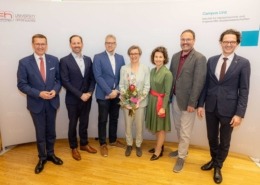 Sieben Personen stehen vor einer Fotowand der FH Oberösterreich am Campus Linz; eine Frau in der Mitte hält einen Blumenstrauß, alle tragen Anzüge.