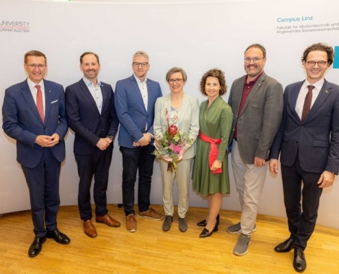Sieben Personen stehen vor einer Fotowand der FH Oberösterreich am Campus Linz; eine Frau in der Mitte hält einen Blumenstrauß, alle tragen Anzüge.