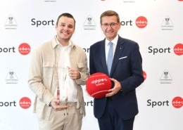 Zwei Herren stehen vor einer weißen Wand mit schriftzug Sport. Ein Mann hält einen Pokal, der andere einen großen Tennisball