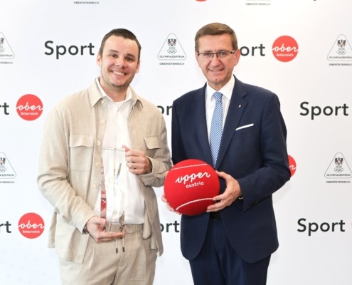 Zwei Herren stehen vor einer weißen Wand mit schriftzug Sport. Ein Mann hält einen Pokal, der andere einen großen Tennisball