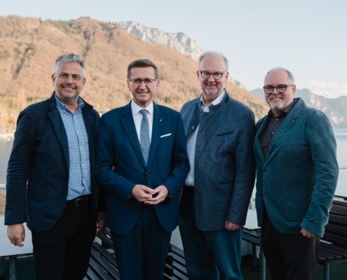 Vier elegant gekleidete Männer stehen vor der Kamera und lächeln. Hinter ihnen ist ein schöner Naturhintergrund.