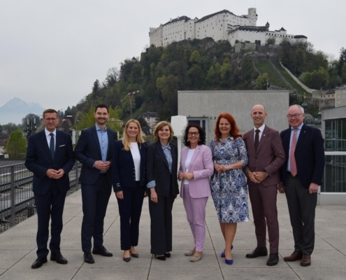 Acht Personen stehen in der Natur in Salzburg und lächeln in die Kamera. Sie sind sehr elegant gekleidet und stehen in einer Linie.