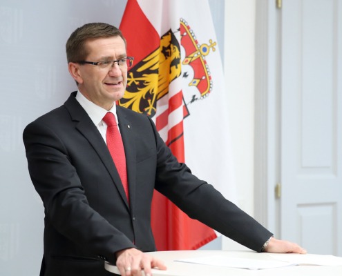 ein Herr steht vor einem Stehtisch und spricht. Hinter ihm ist eine Oberösterreich Flagge.