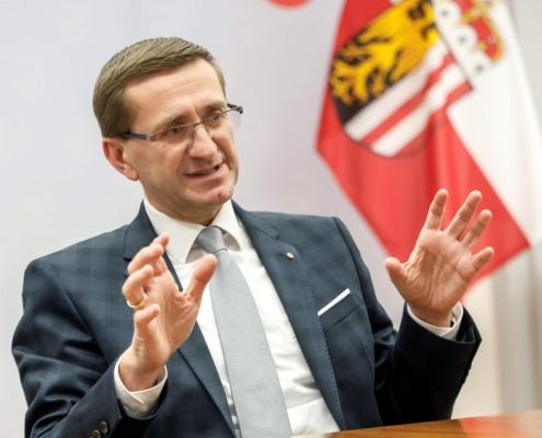 Ein Mann sitz an einem Tisch. Hinter ihm sind OÖ Flaggen.