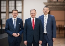 Drei Männer in Anzügen stehen nebeneinander in einem hellen Innenraum und blicken in die Kamera; der Mann in der Mitte trägt eine rote Krawatte, die beiden anderen blaue bzw. graue.
