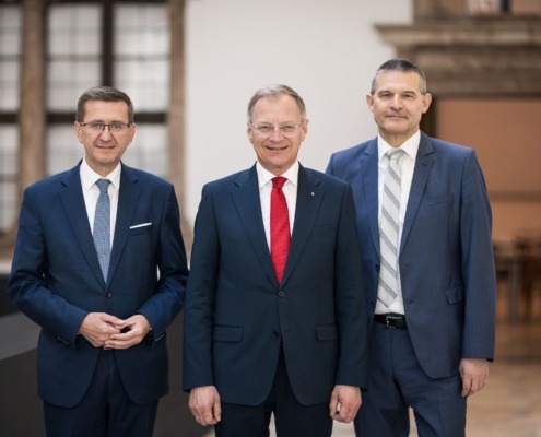 Drei Männer in Anzügen stehen nebeneinander in einem hellen Innenraum und blicken in die Kamera; der Mann in der Mitte trägt eine rote Krawatte, die beiden anderen blaue bzw. graue.