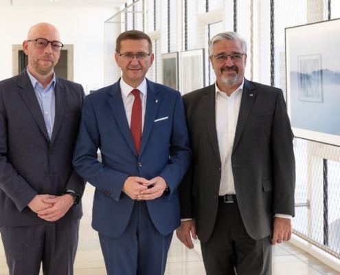 Drei Herren stehen vor der Kamera und lächeln. Sie sind elegant gekleidet.