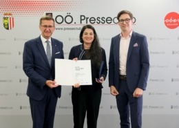Drei Personen stehen elegant gekleidet vor einer Wand und halten ein Zeugnis in der Hand