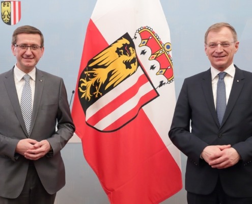 Zwei Herren stehen im Anzug vor einer Wand und lächeln in die Kamera