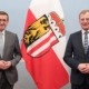 Zwei Herren stehen im Anzug vor einer Wand und lächeln in die Kamera