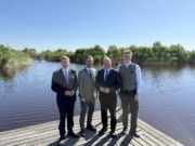 Vier Männer in Anzügen stehen vor einem See und lächeln.