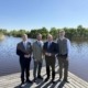 Vier Männer in Anzügen stehen vor einem See und lächeln.