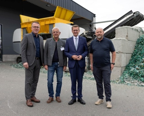 Vier Männer, zwei davon im Anzug, stehen vor einer Recycling-Anlage.