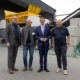 Vier Männer, zwei davon im Anzug, stehen vor einer Recycling-Anlage.