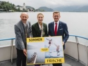 Drei Personen stehen auf einem Boot und halten ein Plakat in der Hand während sie in die Kamera lächeln.