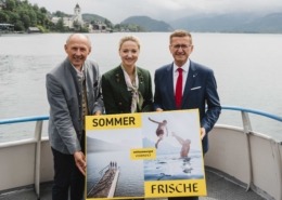 Drei Personen stehen auf einem Boot und halten ein Plakat in der Hand während sie in die Kamera lächeln.