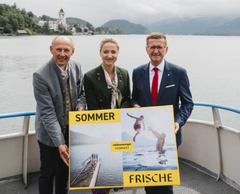 Drei Personen stehen auf einem Boot und halten ein Plakat in der Hand während sie in die Kamera lächeln.