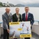 Drei Personen stehen auf einem Boot und halten ein Plakat in der Hand während sie in die Kamera lächeln.