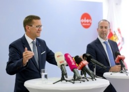 Zwei Herren stehen an einem Tisch mit Mikrofonen und reden.