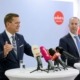 Zwei Herren stehen an einem Tisch mit Mikrofonen und reden.