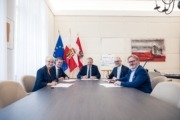 Fünf Personen sitzen an einem Tisch. Hinter ihnen sind zwei Flaggen zu sehen.