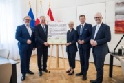 Fünf Personen stehen elegant gekleidet neben einem Poster und lächeln in die Kamera.