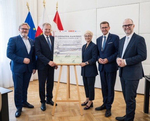 Fünf Personen stehen elegant gekleidet neben einem Poster und lächeln in die Kamera.