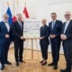 Fünf Personen stehen elegant gekleidet neben einem Poster und lächeln in die Kamera.