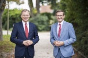 Zwei Herren stehen elegantgekleidet in der Natur und lächeln in die Kamera.