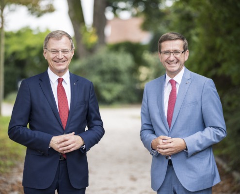 Zwei Herren stehen elegantgekleidet in der Natur und lächeln in die Kamera.