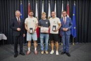 Landeshauptmann Mag. Thomas Stelzer und Wirtschafts- und Sport-Landesrat Markus Achleitner gratulieren der Union Tigers Vöcklabruck. Fünf Herren stehen vor vielen Flaggen und halten Auszeichnungen in der Hand.