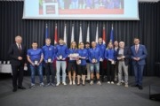 Landeshauptmann Mag. Thomas Stelzer und Wirtschafts- und Sport-Landesrat Markus Achleitner gratulieren dem ASKÖ Traun. Viele Personen in blau angezogen stehen mit Auszeichnungen vor vielen Flaggen.